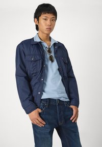 Marineblauw gewatteerd jack met twee borstzakken, gedragen over een lichtblauwe polo. Donkerdenim spijkerbroek met een lichte vervaging en een klassieke snit.