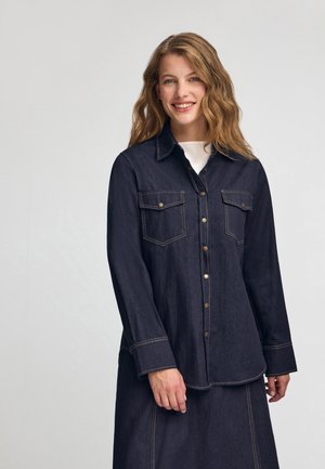 Mørk denimskjorte med lange ærmer, to brystlommer, guldhardware knapper og kontrastbroderi; let krummet kant.