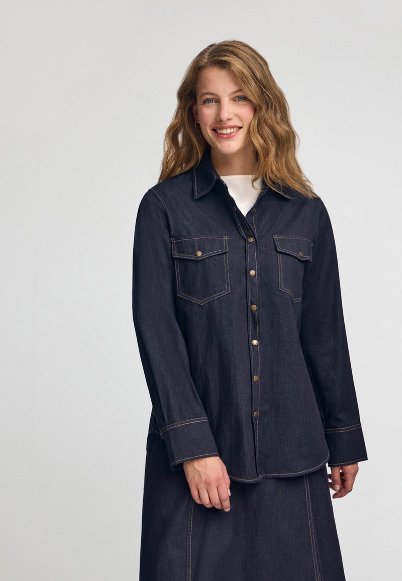 Mørk denimskjorte med lange ærmer, to brystlommer, guldhardware knapper og kontrastbroderi; let krummet kant.
