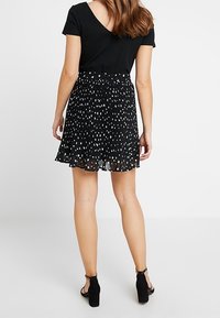 Jupe plissée noire avec des pois blancs, tissu superposé et ourlet festonné. Associée à un haut noir ajusté et des sandales à talons noires.