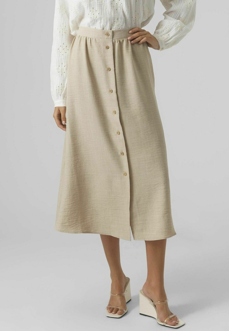Femme portant une jupe midi beige boutonnée devant, un chemisier blanc à manches longues avec des détails en broderie anglaise, et des sandales compensées beige avec des brides transparentes.