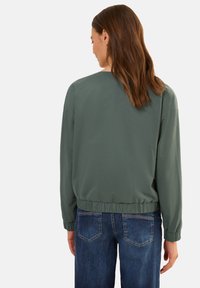 Zelená bombarďácká bunda s elastickým pasem a manžetami, hladký povrch, nošena přes modré džíny. Pohled zezadu, který ukazuje jednoduchý design.
