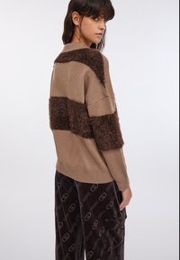 Maglione marrone lavorato a maglia con pannelli in pelliccia sintetica testurizzata, caratterizzato da scollo rotondo e spalle scese. Abbinato a pantaloni scuri con motivo a fantasia.