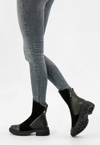 Osoba v šedých úzkých džínách a černých kotníkových botách se zipy po stranách, kráčející na bílém pozadí.