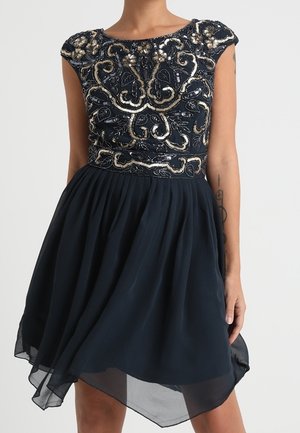 Robe noire sans manches avec un corsage orné de perles dorées et argentées et une jupe en chiffon fluide à ourlet asymétrique portée par une personne debout.