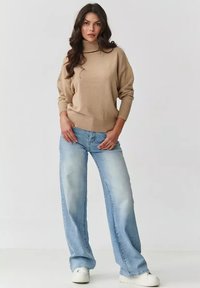 Beżowy, oversize sweter z golfem z prążkowanym wykończeniem, zestawiony z jasnoniebieskimi, szerokimi dżinsami. Białe trampki dopełniają stylizację.