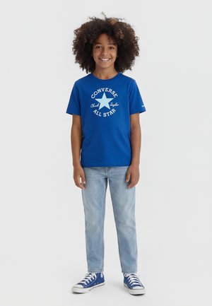 Lächelndes Kind mit lockigem Haar, das ein blaues Converse All Star T-Shirt, helle Jeans und blaue High-Top-Sneaker trägt, steht vor weißem Hintergrund.