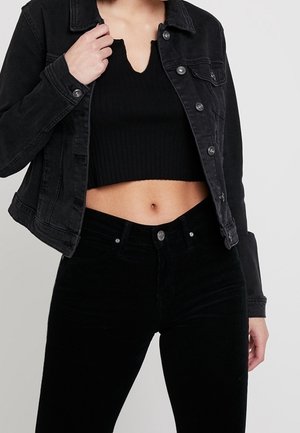 Frau trägt ein schwarzes, kurzes, geripptes Top, schwarze Denimjacke und hoch taillierte schwarze Hose, eine Hand hält den Jackenkragen.