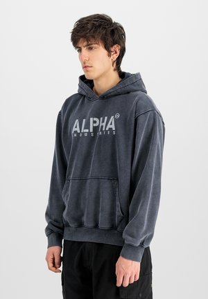 Young man with dark hair wearing a faded black "Alpha Industries" hoodie and black pants, standing against a plain light background.
