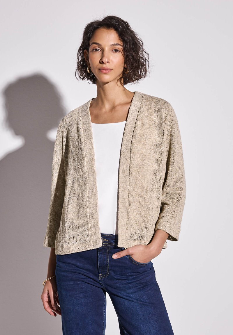 Beige stickad cardigan med öppen front, trekvartslånga ärmar och en texturerad yta, kombinerad med en vit topp och mörkblå jeans.
