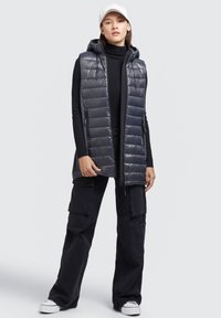 Graue Steppweste mit Kapuze, mit quilted Design und mit Reißverschlüssen versehenen Taschen, getragen über einem schwarzen Rollkragenpullover, kombiniert mit schwarzen Cargo-Hosen.