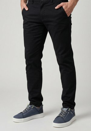 Mann trägt schwarze eng anliegende Hose und marineblaue Sneaker mit weißen Sohlen, steht mit den Händen in den Taschen vor neutralem Hintergrund.