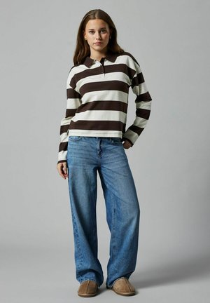 Langærmet stribet polo-shirt i brun og creme, parret med brede lyseblå jeans og brune slippers, stående mod en grå baggrund.