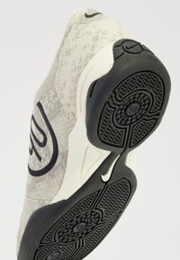 Zapatilla de deporte gris claro con parte superior texturizada que presenta un patrón de piel de serpiente, acentos negros y una suela de goma estampada. Logo de Nike visible.