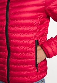 Červená vycpaná bunda s horizontálním prošíváním, s černým zipem a detailem kapes. Hladká textura a přiléhavé manžety.