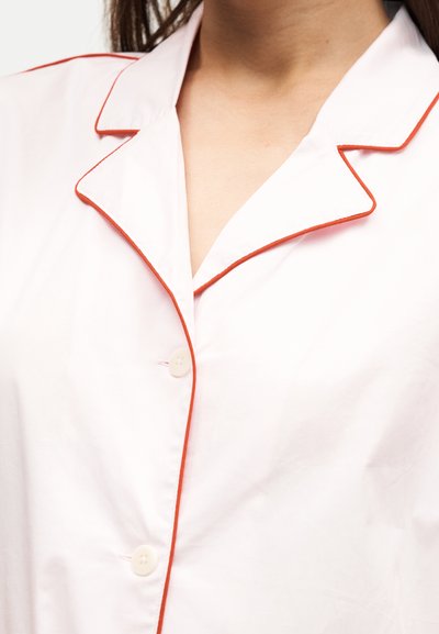 Gros plan d'une chemise blanche avec un passepoil rouge sur le col et la ligne de boutons, montrant deux boutons blancs attachés.