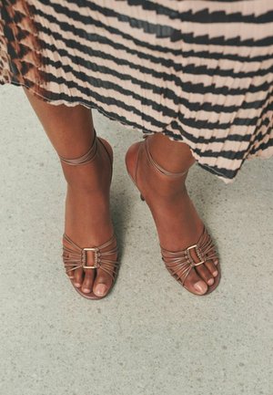 Füße in braunen Riemchensandalen mit Knöchelriemen, stehend auf einem hellgrauen strukturierten Boden, mit einem schwarz-beigen gestreiften plissierten Rock.
