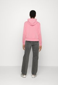 Rosa kort hoodie med snörning i huvan, kombinerad med löst sittande grå jeans och flerfärgade sneakers, sedd bakifrån.