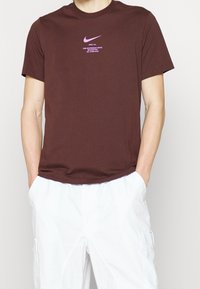 Brązowy bawełniany t-shirt z fioletowym logo Nike i grafika tekstową na klatce piersiowej. Krótkie rękawy i luźny krój. Połączony z białymi szortami.