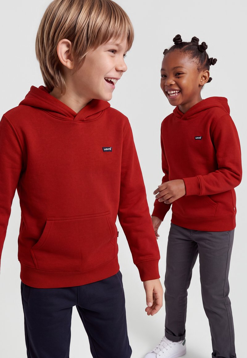 Deux enfants riant et debout, tous deux portant des sweats à capuche rouges Levi's, pantalons foncés et baskets sur un fond clair uni.