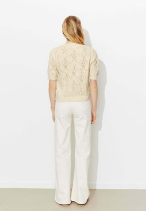 Femme aux longs cheveux blonds debout face à un mur blanc, portant un haut beige en maille à manches courtes et un pantalon blanc à jambes larges.