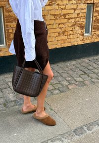 Borsa a spalla in pelle intrecciata marrone con due manici, abbinata a una gonna lavorata a maglia scura e scarpe slip-on in pelle di volpe marroni, su una strada lastricata.