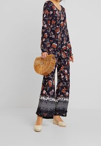 Kvinde iført en mørk blomsterprint langærmet top og bukser, der holder en træ halvmåne håndtaske, iført beige kile-espadriller.
