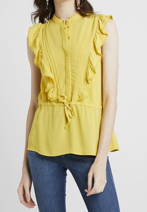 Kvinde iført ærmeløs gul bluse med flæser foran og bindedetalje, kombineret med blå jeans mod en hvid baggrund.