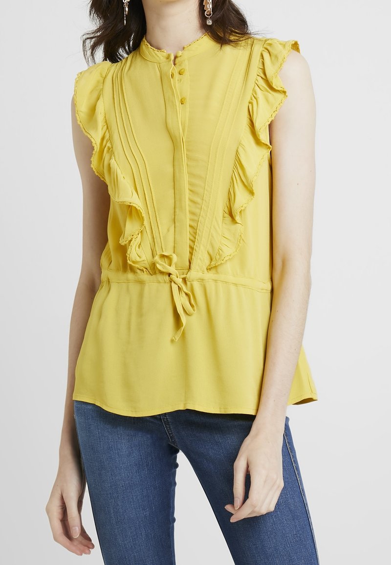 Femme portant un chemisier sans manches jaune avec des volants à l'avant et un détail à nouer, associé à un jean bleu sur un fond blanc.