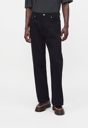 Personne portant un jean noir droit, des chaussures en cuir marron foncé avec un détail chaîne, et une chemise à manches courtes gris foncé.