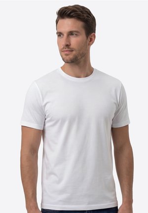 Man met kort bruin haar die een eenvoudige witte T-shirt met korte mouwen en ronde hals draagt, kijkt iets naar links tegen een effen achtergrond.