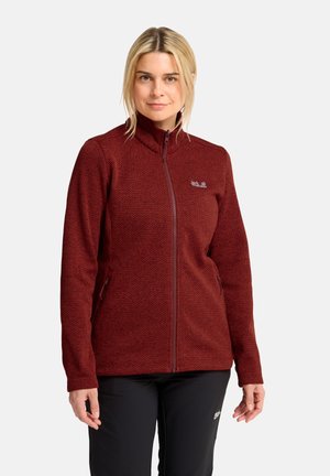 Donna con capelli biondi che indossa una giacca bordeaux con zip e una trama, e pantaloni neri, in piedi su uno sfondo bianco uniforme.