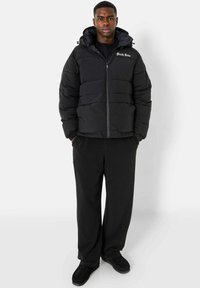 Zwarte gewatteerde jas met ritssluiting en capuchon, met een logo op de borst. Gecombineerd met losse zwarte broek en schoenen.