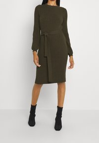 Abito in maglia a coste di un verde oliva scuro, con scollatura rotonda, maniche lunghe, cintura in vita e orlo a lunghezza ginocchio. Abbinato a stivaletti neri alla caviglia.