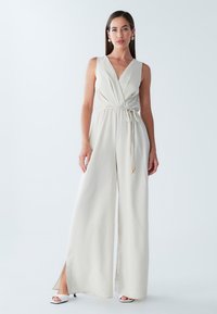 Beige ærmeløs jumpsuit med V-hals, indsnøret talje, bred ben-design, sideslids og snøredetalje. Kombineret med hvide hæle.