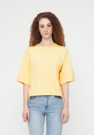 Haut jaune oversize à manches courtes en tissu doux, avec un col rond et un ourlet côtelé, porté avec un jean bleu clair.