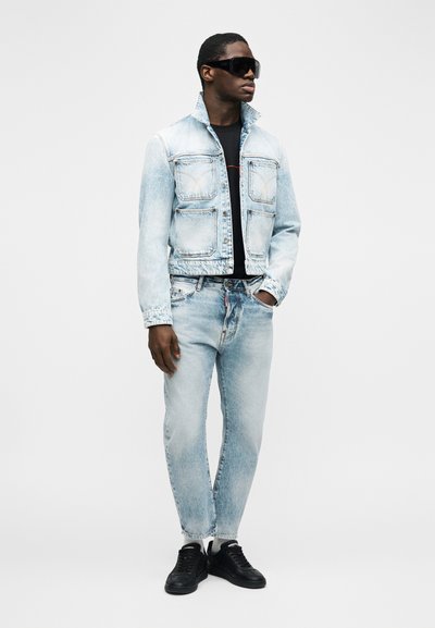 Άνδρας που φοράει ανοιχτό μπλε τζιν μπουφάν και τζιν παντελόνι, μαύρο μπλουζάκι, μαύρα αθλητικά παπούτσια και μεγάλα μαύρα γυαλιά ηλίου, στέκεται με το ένα χέρι στην τσέπη.