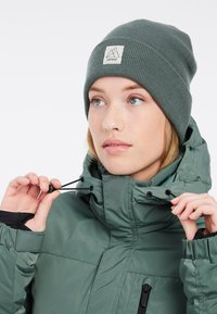 Groene pufferjack met een hoge kraag en een verstelbare trekkoord. Draagt een geribde groene beanie met een vierkant logo-label.