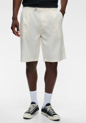 Homme portant un short écru mi-cuisses, des baskets basses noires avec des lacets blancs, des chaussettes blanches et un t-shirt noir sur un fond uni.