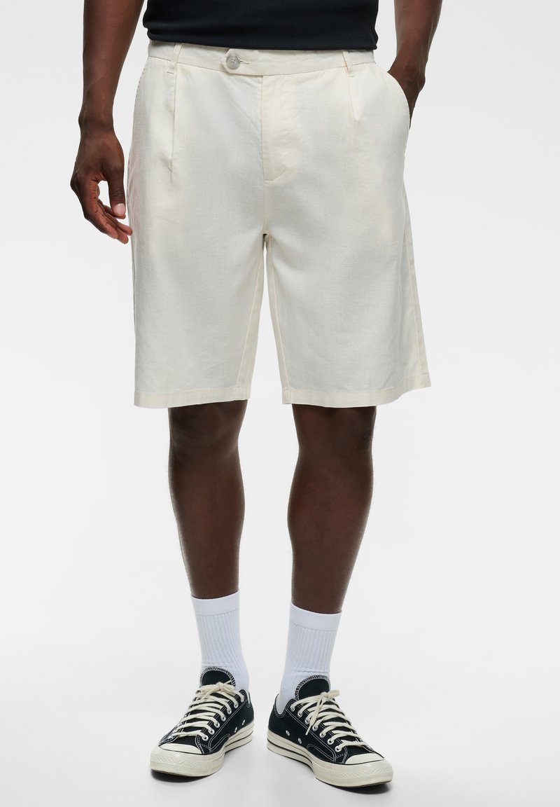 Homme portant un short écru mi-cuisses, des baskets basses noires avec des lacets blancs, des chaussettes blanches et un t-shirt noir sur un fond uni.