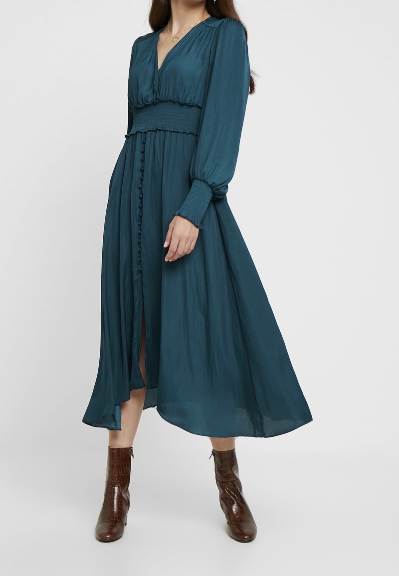 Tealfärgad långärmad klänning med V-ringning, smockad midja, knappstängning framtill och en svävande kjol. Tillsammans med bruna ankelstövlar.