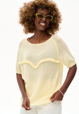 Uśmiechnięta kobieta z kręconymi włosami, nosząca żółte okulary przeciwsłoneczne, jasny żółty top z falbankami i krótkim rękawem, białe spodnie oraz bransoletkę na nadgarstku.