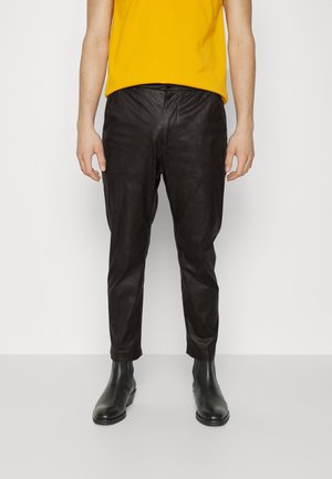 Mann trägt schwarze Lederhose, schwarze Knöchelstiefel und ein gelbes Hemd, steht vor einem schlichten weißen Hintergrund.