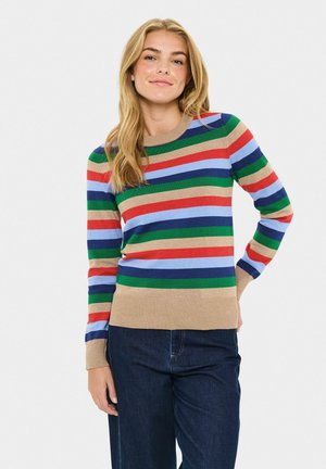 Femme blonde portant un pull à rayures horizontales multicolores et un jean bleu foncé, se tenant avec une main sur la hanche.