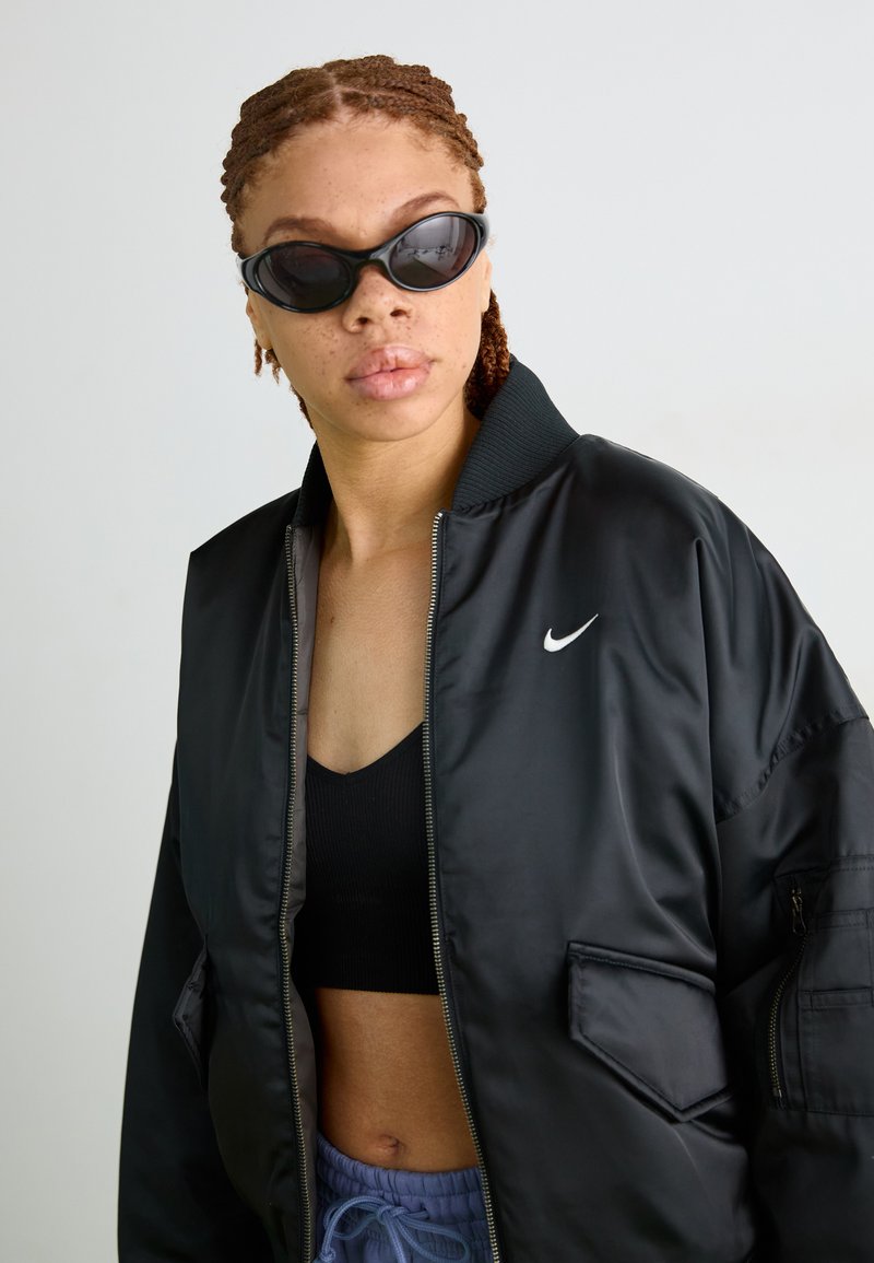Veste bomber noire avec fermeture éclair à l'avant, col côtelé et logo Nike. Portée par-dessus un crop top noir, associée à un pantalon bleu à cordon de serrage.