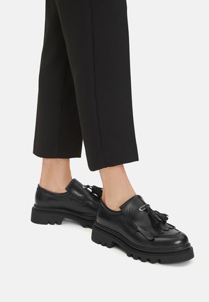 Czarne skórzane buty z masywną podeszwą, ozdobione chwostem i zaokrąglonym kształtem. Połączone z czarnymi, skróconymi spodniami.