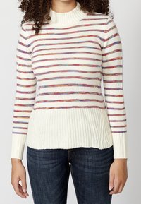 Pull rayé avec un col montant côtelé, présentant des rayures multicolores sur un fond blanc, et un ourlet côtelé. Associé à un jean foncé.