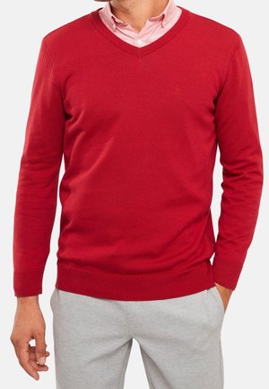Mann trägt roten V-Ausschnitt-Pullover über einem hellrosa Hemd mit hellgrauer Hose, Hände entspannt an den Seiten, Kopf nicht sichtbar.