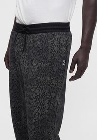 Άνδρας φοράει μαύρο παντελόνι φόρμας Emporio Armani με επαναλαμβανόμενο τύπωμα λογότυπου και μικρή ετικέτα EA7 στην τσέπη, σε συνδυασμό με λευκό πουκάμισο.