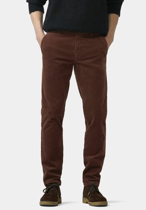 Pantalon en velours côtelé marron à coupe slim, avec poches avant et texture lisse, associé à des chaussures en daim foncé.